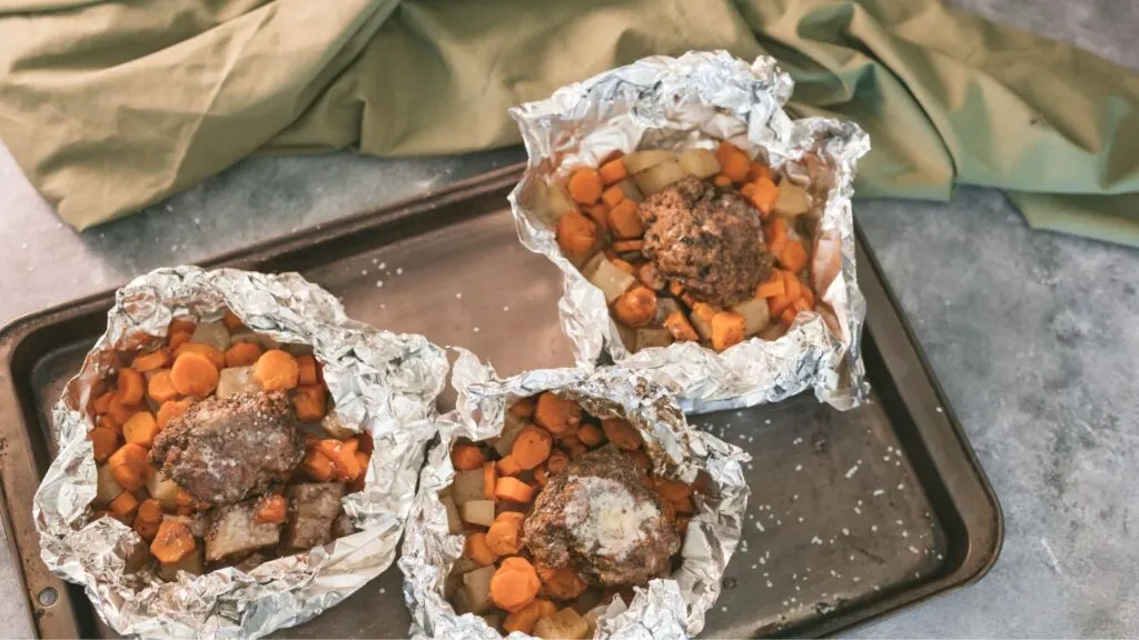 Three hobo dinner foil packets with hamburger patty, carrots, potatoes and onions arranged on a sheet pan.