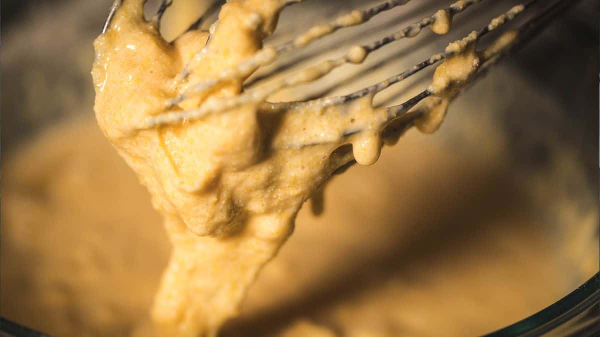 A close-up shot of thick cornbread batter clinging to a whisk, showing its creamy and textured consistency. The blurred background emphasizes the batter's rich, golden color, evoking the warmth of homemade baking.