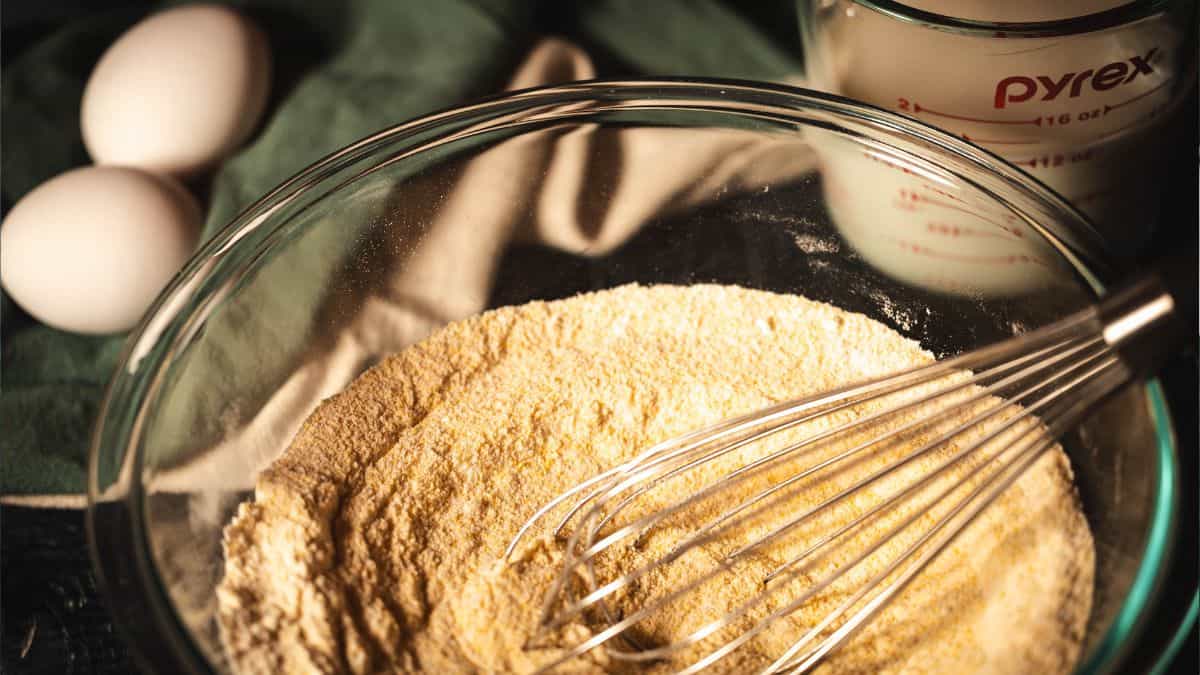 A glass mixing bowl filled with dry cornbread ingredients, including cornmeal and flour, with a whisk resting inside. Two eggs and a measuring cup of milk sit in the background on a soft green and cream cloth, illuminated by warm light, evoking a cozy baking scene.