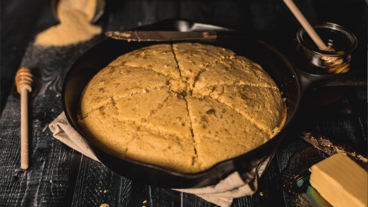 A golden-brown cornbread baked in a cast-iron skillet, sliced into wedges and resting on a neutral cloth. A honey dipper, a butter knife with crumbs, and a pat of butter on a decorative plate sit nearby on a dark wooden surface, enhancing the rustic and cozy presentation.