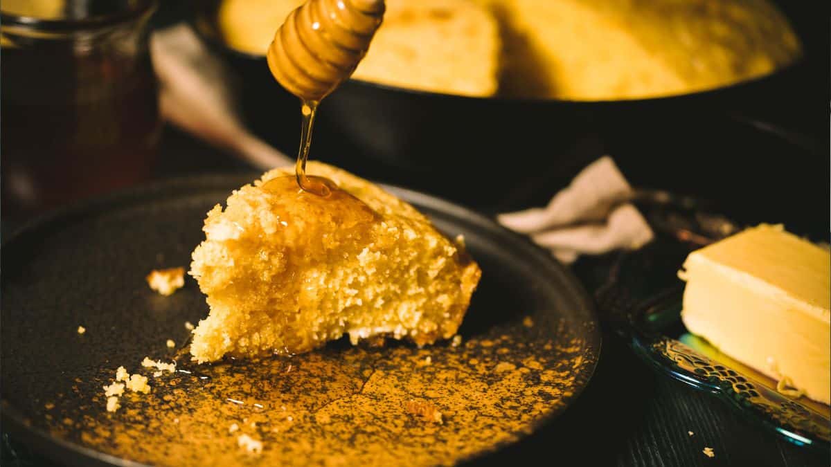 A close-up shot of a golden slice of cornbread on a dark plate, with honey being drizzled over the top. Crumbs are scattered around, and a pat of butter sits on a decorative plate in the background, creating a warm and appetizing scene.