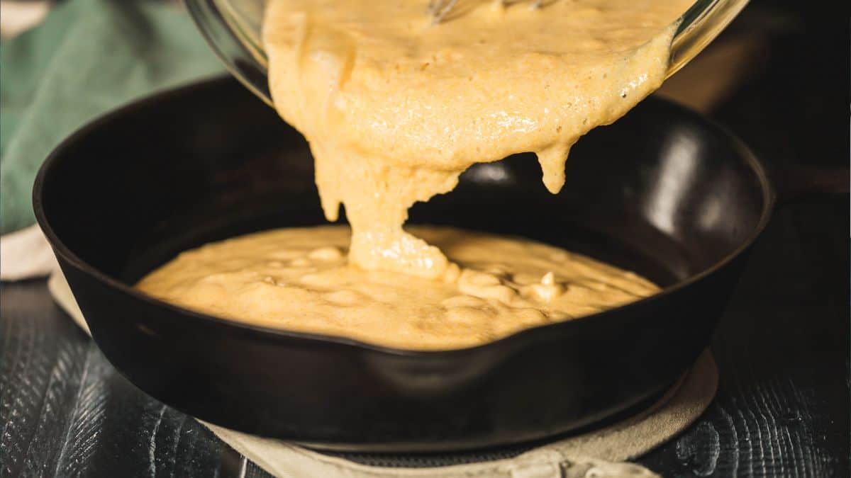 A close-up action shot of cornbread batter being poured from a glass bowl into a preheated cast-iron skillet. The batter's smooth and airy texture is highlighted against the dark skillet, evoking the start of a homemade baking process.