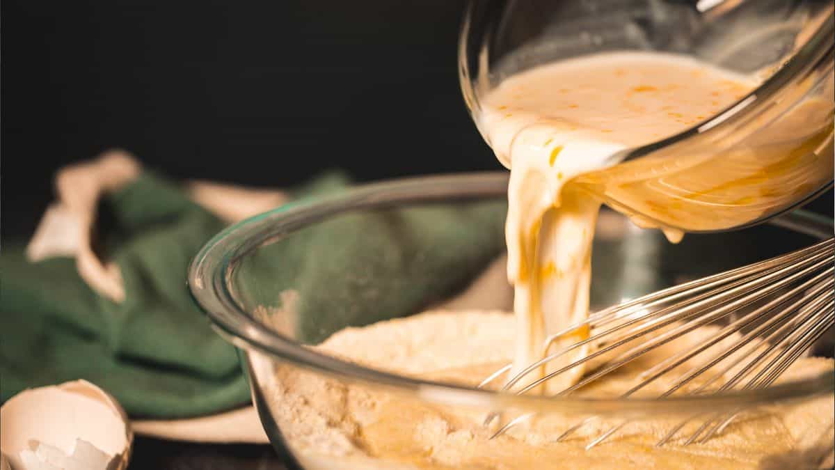 A close-up action shot of wet ingredients, including milk and eggs, being poured from a glass bowl into a mixing bowl with dry cornbread ingredients. A whisk rests in the bowl, and a cracked eggshell and green cloth are blurred in the background, adding a rustic, homemade feel.