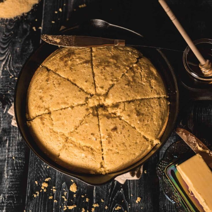 A rustic skillet cornbread sliced into wedges, showcasing its golden crust and soft texture. The cornbread is served in a cast-iron skillet, surrounded by butter, a honey jar, and scattered crumbs on a dark wood surface, creating a warm and inviting homemade feel.