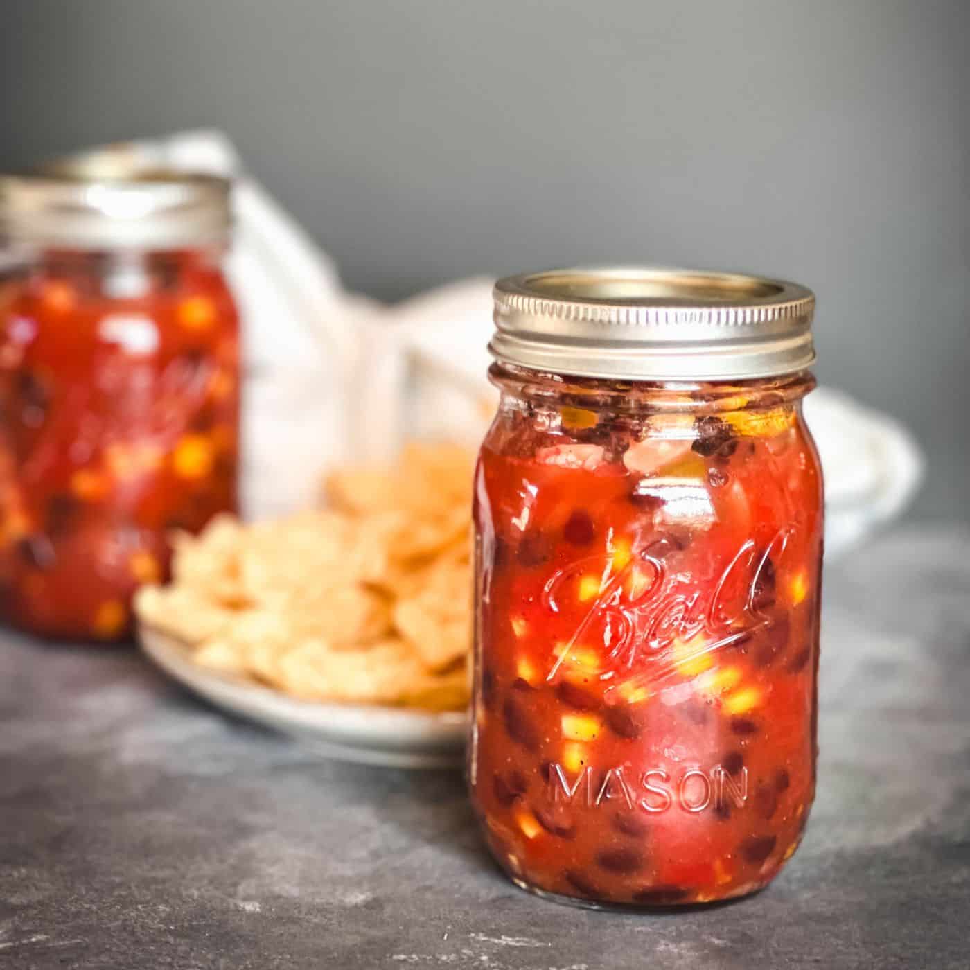 Mason jars full of black bean and corn salsa with tortilla chips in the background