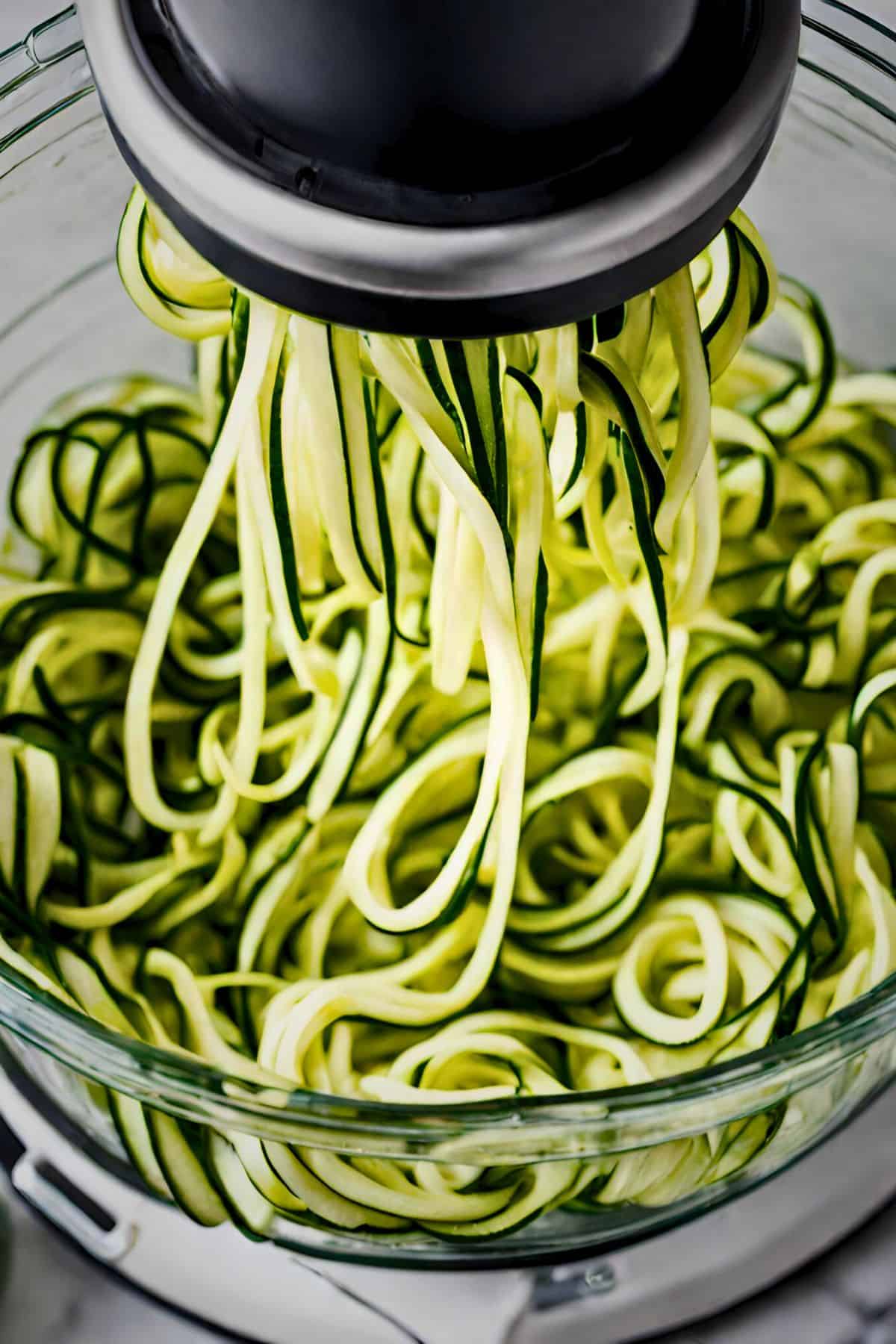 Zucchini being spiralized into zoodles using a countertop spiralizer, with fresh green noodles falling into a glass bowl below.
Zucchini being spiralized into zoodles using a countertop spiralizer, with fresh green noodles falling into a glass bowl below.