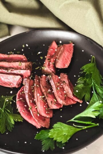 Pan-fried deer heart slices garnished with fresh parsley on a black plate, showcasing a rustic and adventurous dish.