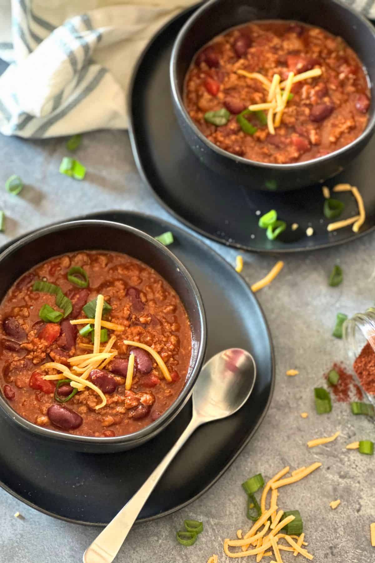 Two bowls of homemade venison chili with beans, topped with shredded cheese and green onions, served with a spoon on a black plate.