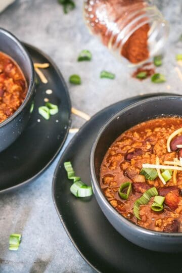 Bowl of hearty venison chili with kidney beans, topped with shredded cheese and green onions, served in a black bowl on a dark plate.