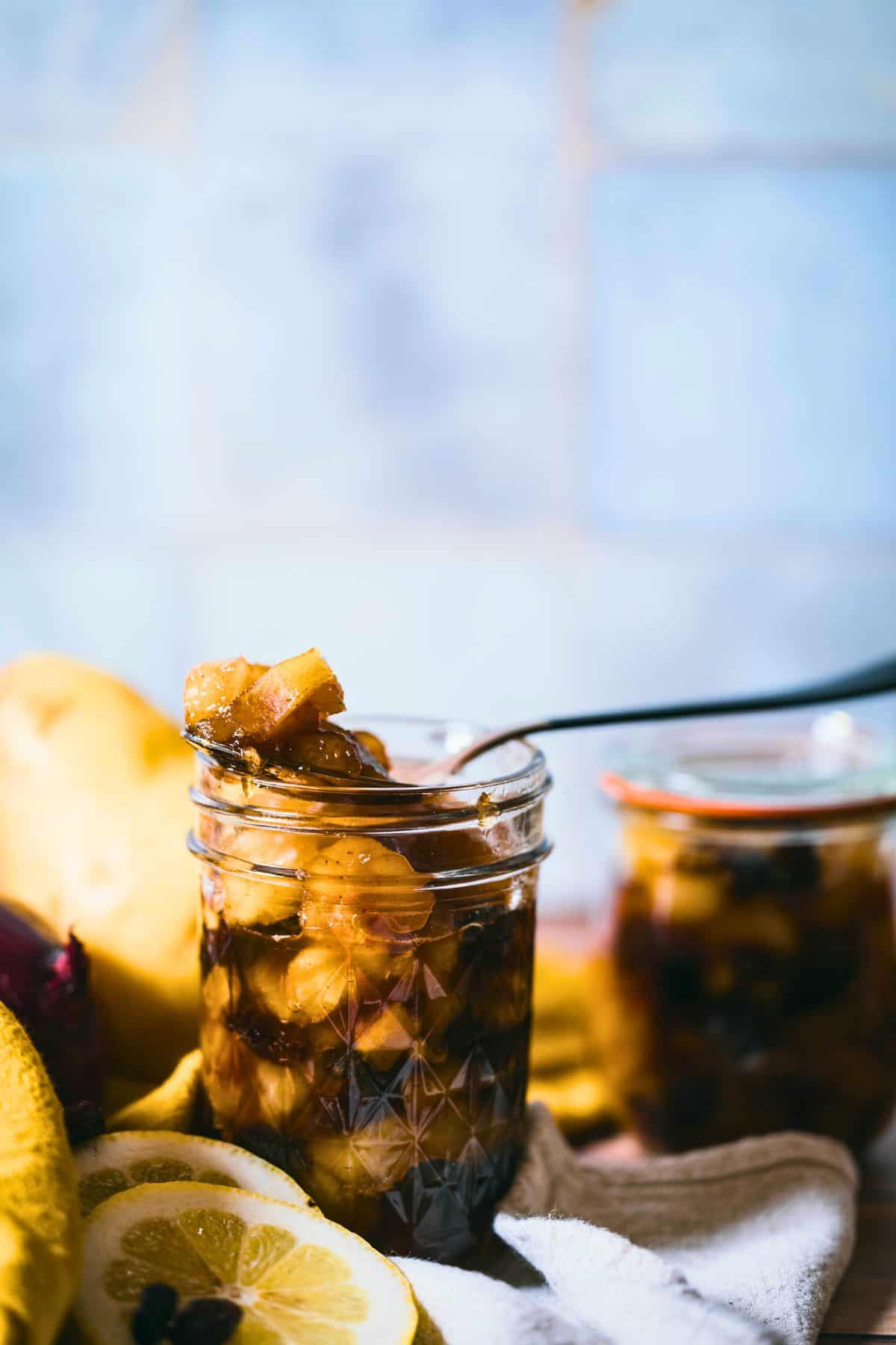 Jar of homemade spiced pear chutney with a spoonful lifted above, showing tender pears, raisins, and warm spices, with fresh lemon slices and another jar blurred in the background.