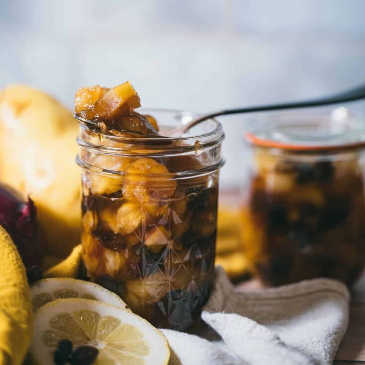 Jar of homemade spiced pear chutney with a spoonful lifted above, showing tender pears, raisins, and warm spices, with fresh lemon slices and another jar blurred in the background.
