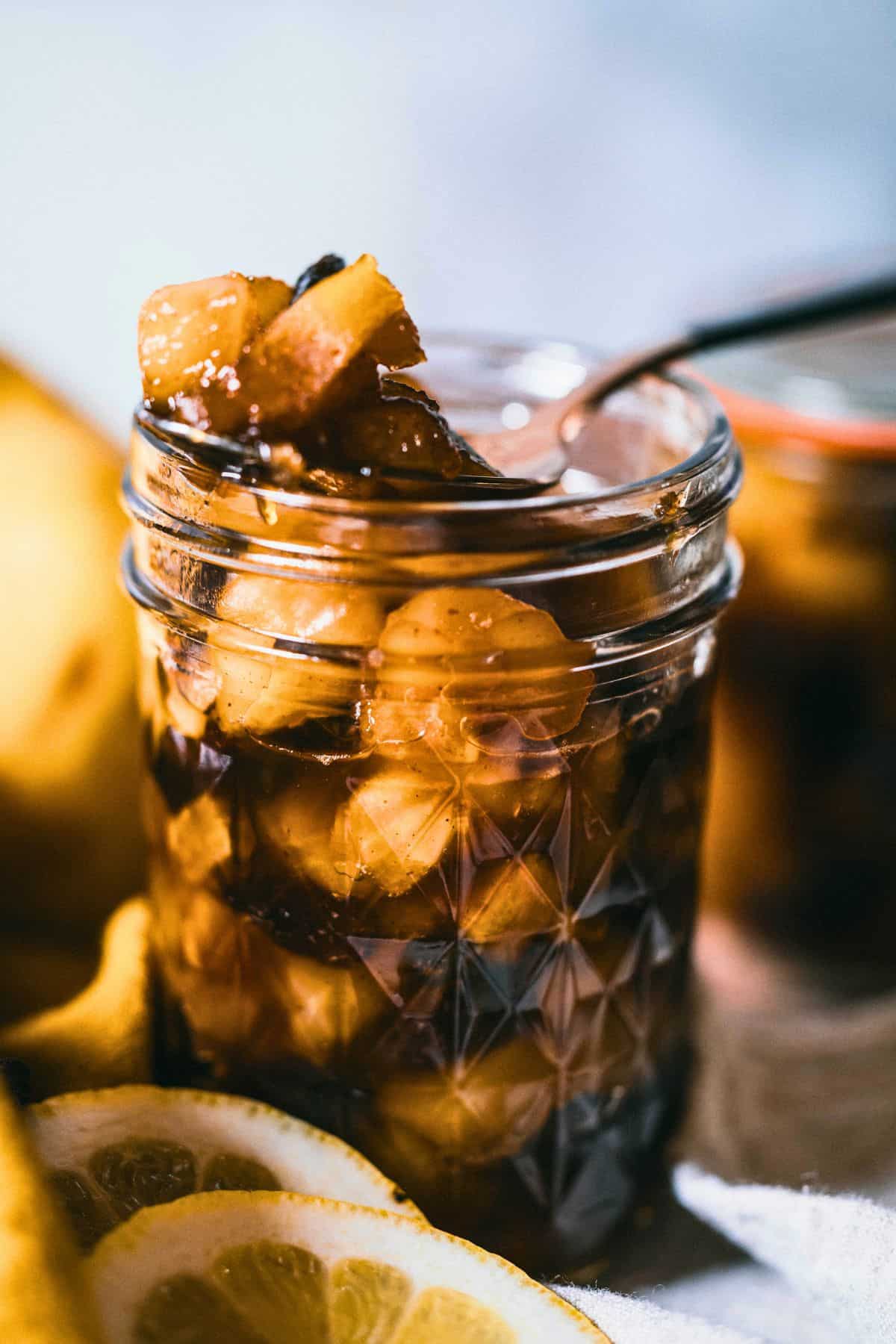Close-up of homemade spiced pear chutney in a jar with a spoon lifting tender pear pieces, showing the thick texture and warm spices, with lemon slices blurred in the foreground.