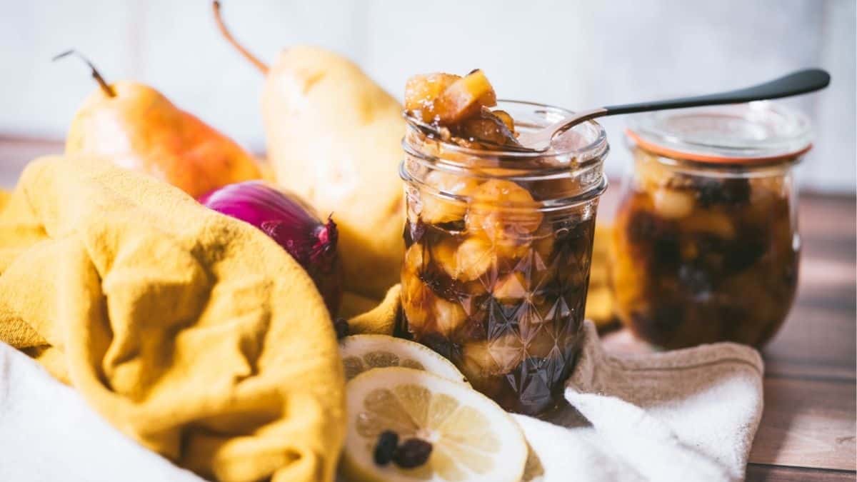 Landscape photo of homemade spiced pear chutney in an open jar with a spoon, surrounded by fresh pears, lemon slices, raisins, and a second jar in the background.