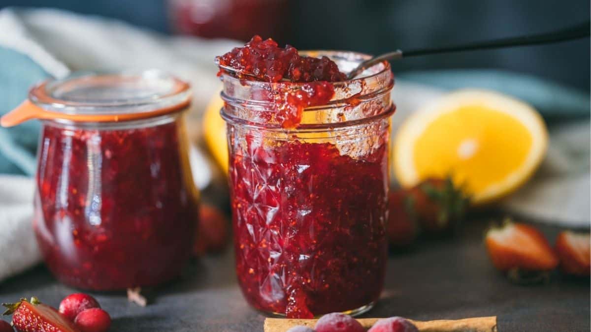 Homemade cranberry strawberry Christmas jam in glass jars with oranges and spices in the background, perfect for gifting during the holidays.