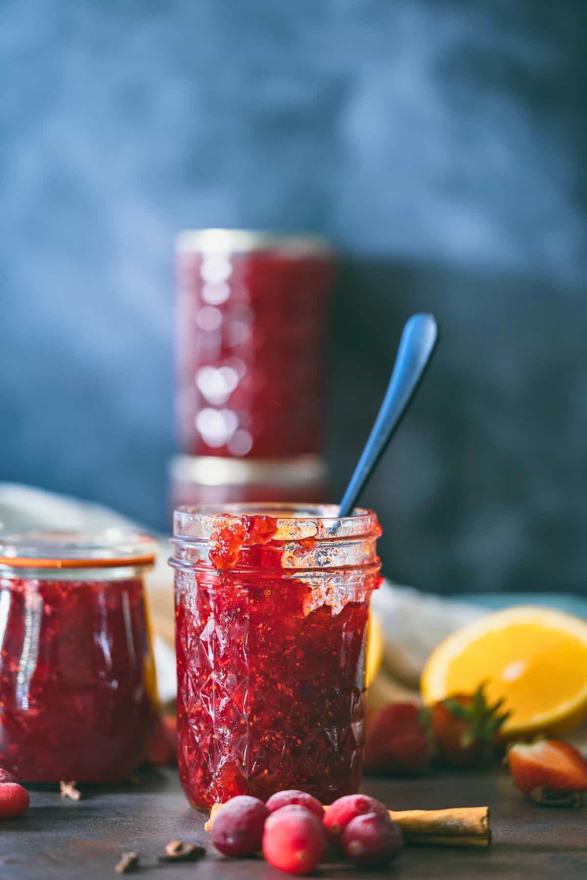 Glass jar filled with homemade Christmas jam made from cranberries, strawberries, and oranges, surrounded by fresh fruit and a cinnamon stick on a dark rustic surface.