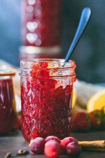 Glass jar filled with homemade Christmas jam made from cranberries, strawberries, and oranges, surrounded by fresh fruit and a cinnamon stick on a dark rustic surface.