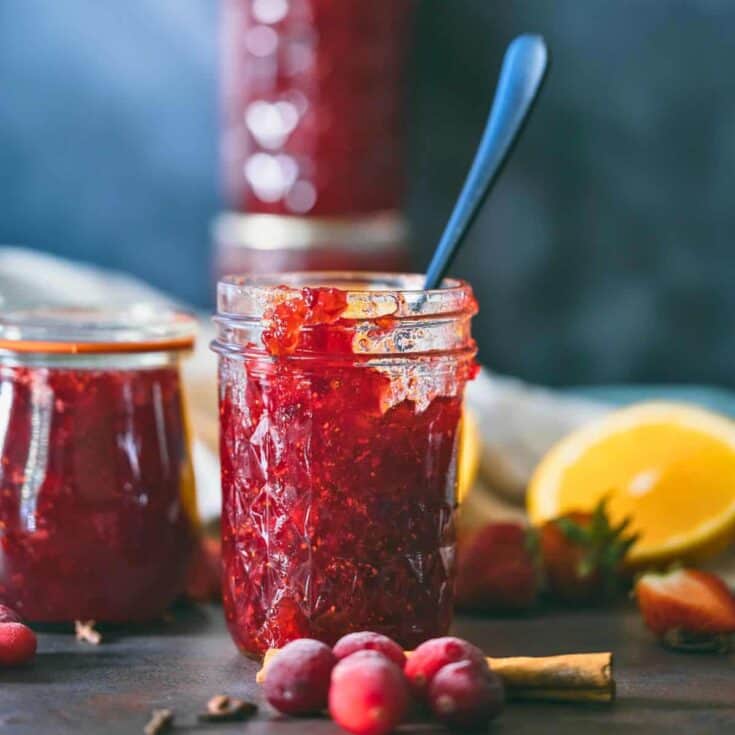 Glass jar filled with homemade Christmas jam made from cranberries, strawberries, and oranges, surrounded by fresh fruit and a cinnamon stick on a dark rustic surface.