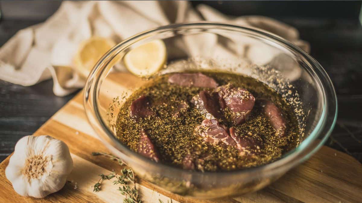 Venison medallions marinating in a glass bowl with olive oil, herbs, garlic, and lemon on a wooden cutting board.