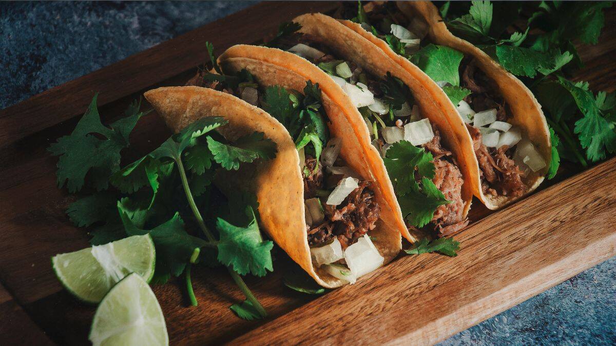 Street-style venison tacos topped with chopped onions and fresh cilantro, served on a wooden board with lime wedges.