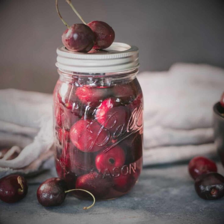 Close-up of a mason jar filled with homemade brandied cherries, rich red fruit soaked in syrup, with fresh cherries scattered around on a rustic surface—perfect garnish for cocktails or desserts.