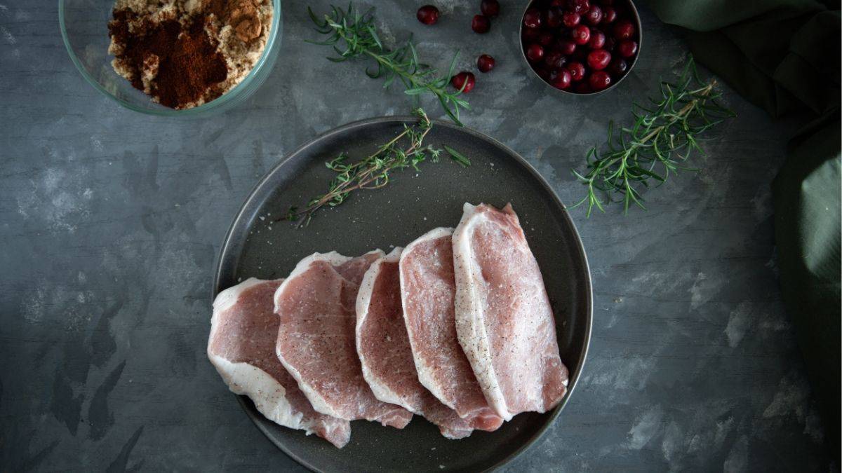 Raw pork chops on a plate surrounded by fresh cranberries, rosemary, thyme, and a bowl of brown sugar chili rub for making cranberry pork chops.