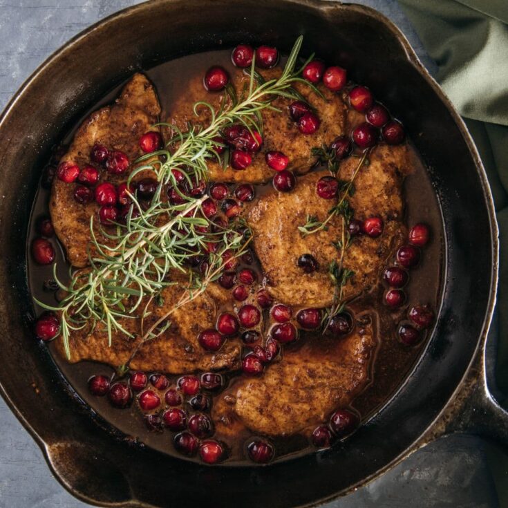 Cast iron skillet filled with seared pork chops simmered in a cranberry and red wine sauce, topped with fresh rosemary and thyme.