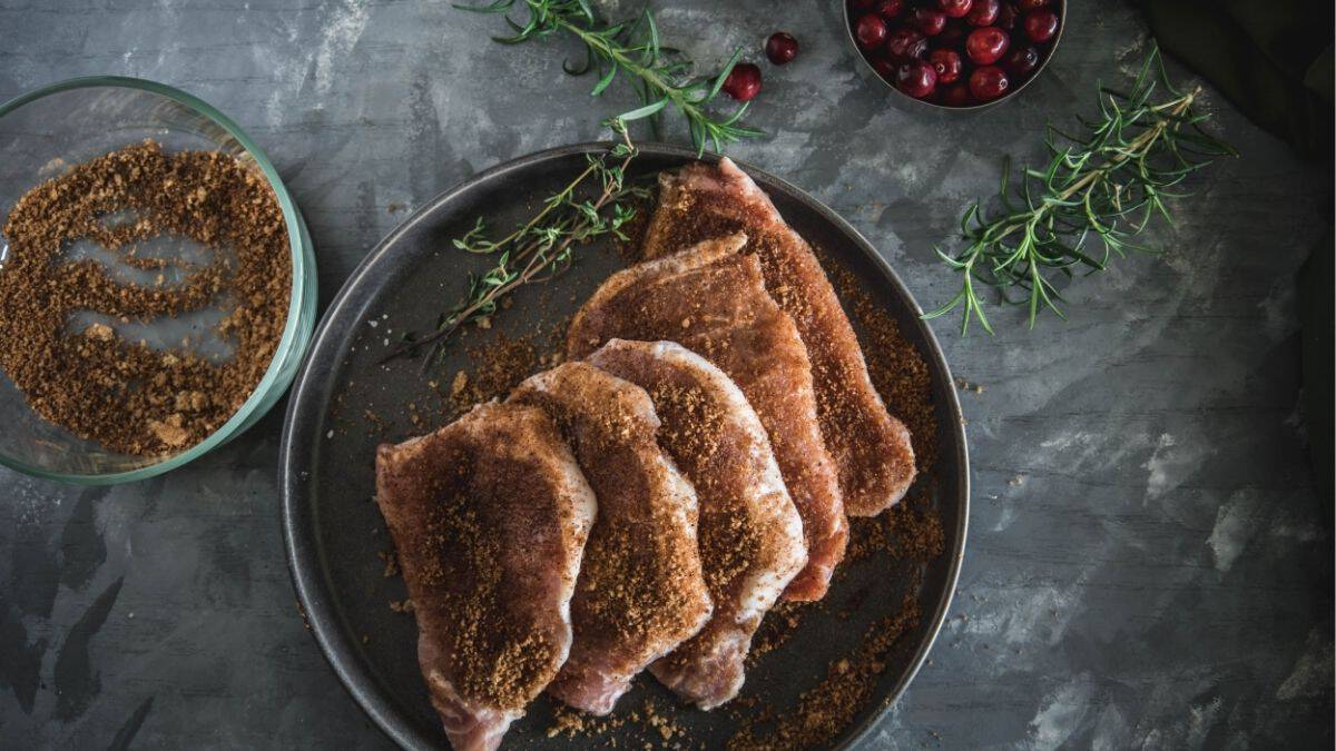 Pork chops coated in a brown sugar and chili powder rub on a plate, with fresh rosemary, thyme, cranberries, and a bowl of the spice mixture beside them.