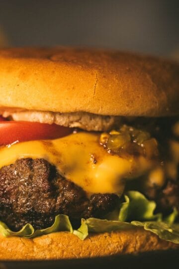 Close-up of a juicy grilled burger with melted cheddar, tomato slices, lettuce, and relish on a toasted bun.