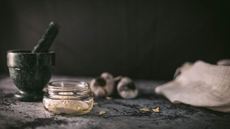 Jar of garlic powder on a counter surrounded by dried garlic, fresh garlic, and a mortar & pestle.