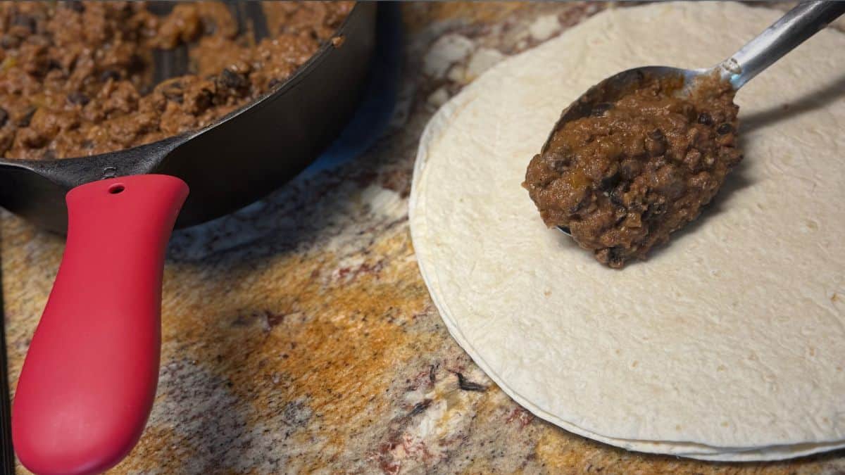 Ground venison and bean mixture being spooned onto a flour tortilla next to a cast iron skillet on a stone countertop.