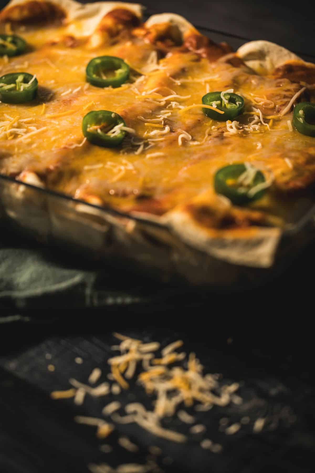 Baked venison enchiladas topped with melted cheese and sliced jalapeños in a glass baking dish, with shredded cheese scattered on a dark surface.