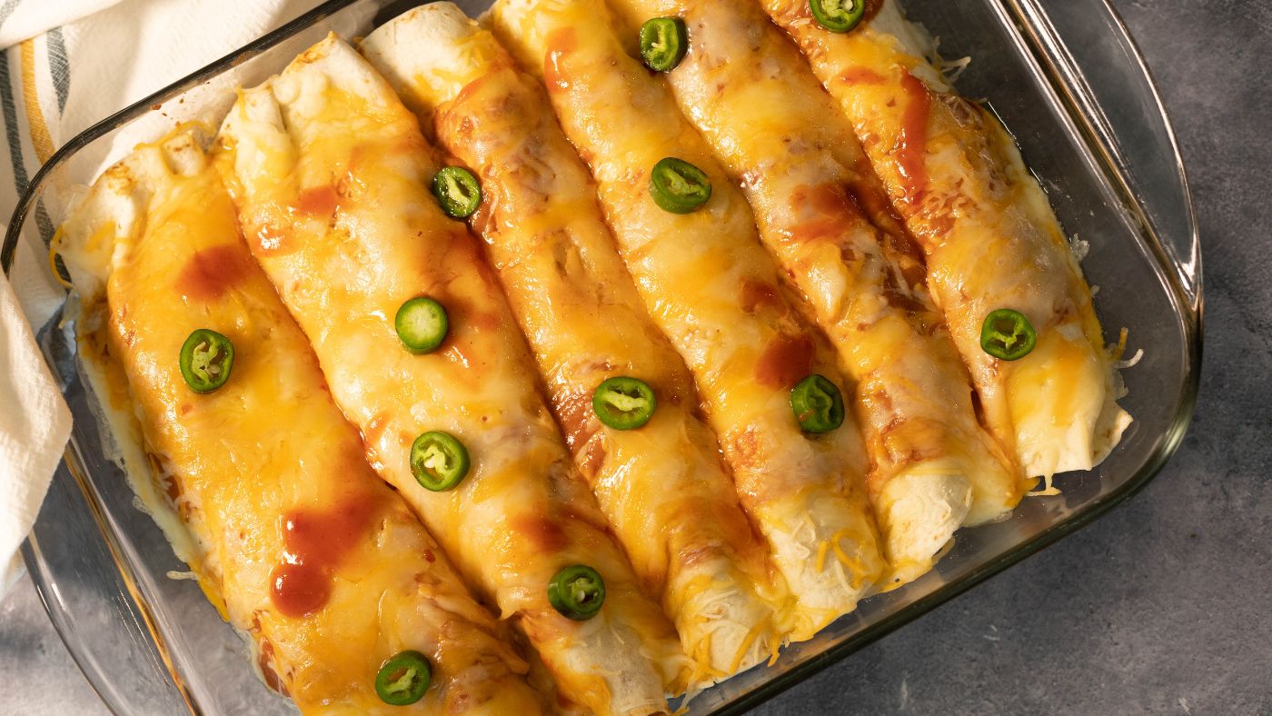 A dish of venison enchiladas covered in cheese and fresh jalapeños on a dark countertop.