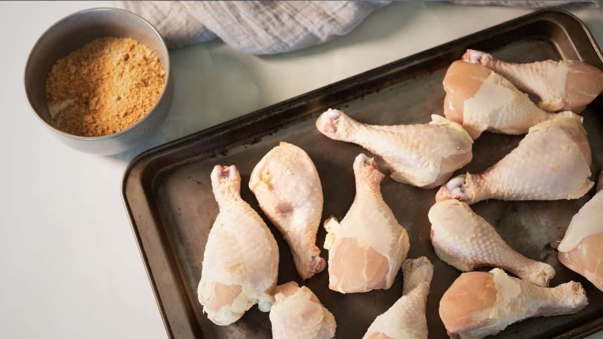 Raw chicken drumsticks arranged on a baking sheet next to a bowl of dry rub seasoning, ready for smoking.