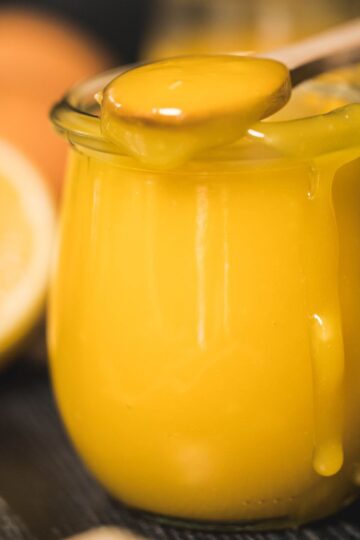 A close-up of a jar filled with silky, golden lemon curd, with a wooden spoon resting on top, dripping with the rich, smooth curd. A second jar, fresh lemons, and eggs blur softly in the background, creating a warm, moody, and inviting kitchen scene.
