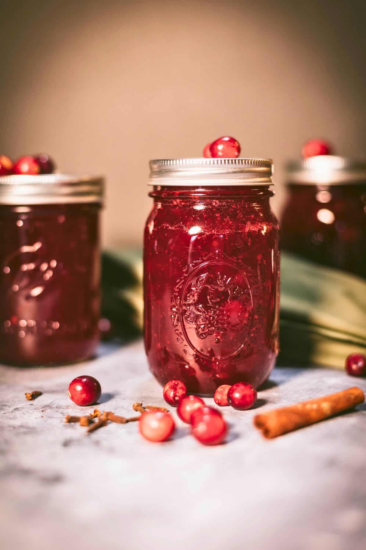 Jars of homemade cranberry sauce ready for canning with fresh cranberries, cloves, and cinnamon on the counter