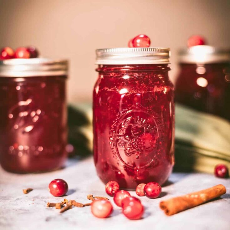 Jars of homemade cranberry sauce ready for canning with fresh cranberries, cloves, and cinnamon on the counter.