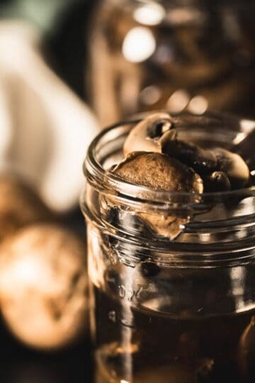 A glass jar filled with pressure-canned mini bella mushrooms, with a fork lifting mushrooms from the jar; whole mushrooms and jars blurred in the background.
