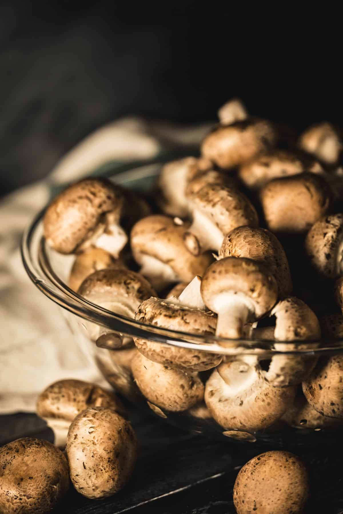 A glass bowl filled with fresh mini bella mushrooms, with a few loose mushrooms scattered on a dark surface in front of a light cloth.
