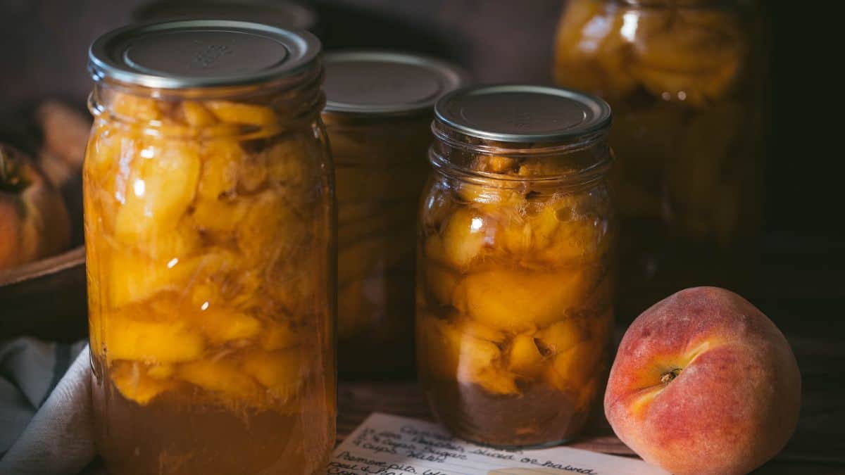 Jars of homemade canned peaches in light syrup beside a fresh peach and handwritten recipe card after water bath canning.
