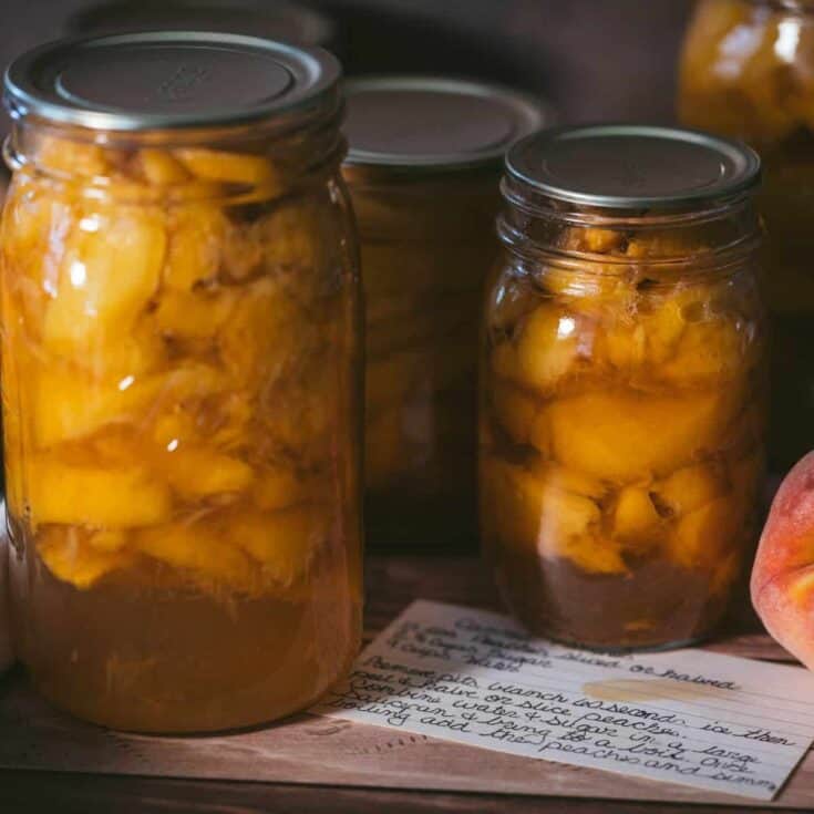 Jars of homemade canned peaches in light syrup beside a fresh peach and handwritten recipe card after water bath canning.