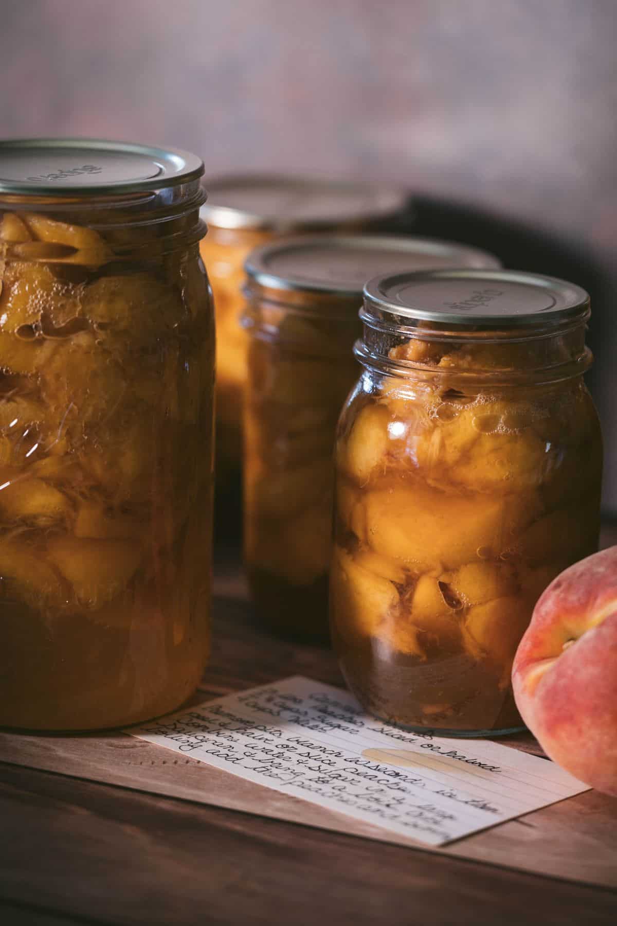 Jars of homemade canned peaches in light syrup with a fresh peach and handwritten recipe card on a wooden table.
