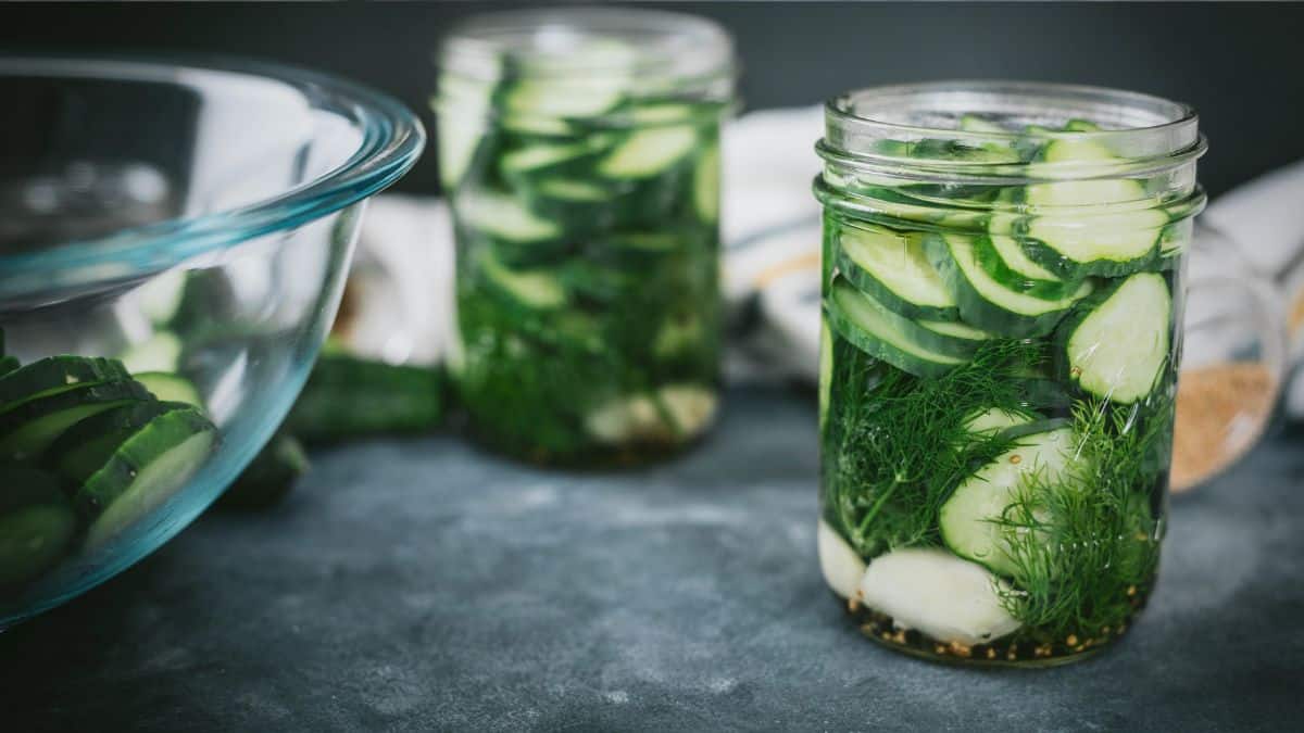 Fresh dill pickles packed into glass jars with garlic and herbs, ready for the refrigerator, with a bowl of sliced cucumbers nearby on a dark countertop