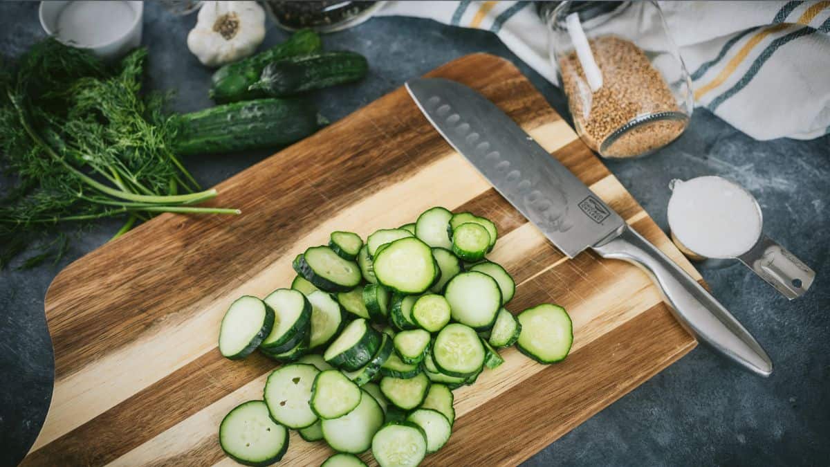 Fresh cucumbers sliced into rounds on a wooden cutting board with a chef’s knife, surrounded by dill, garlic, spices, and salt for making homemade dill pickles