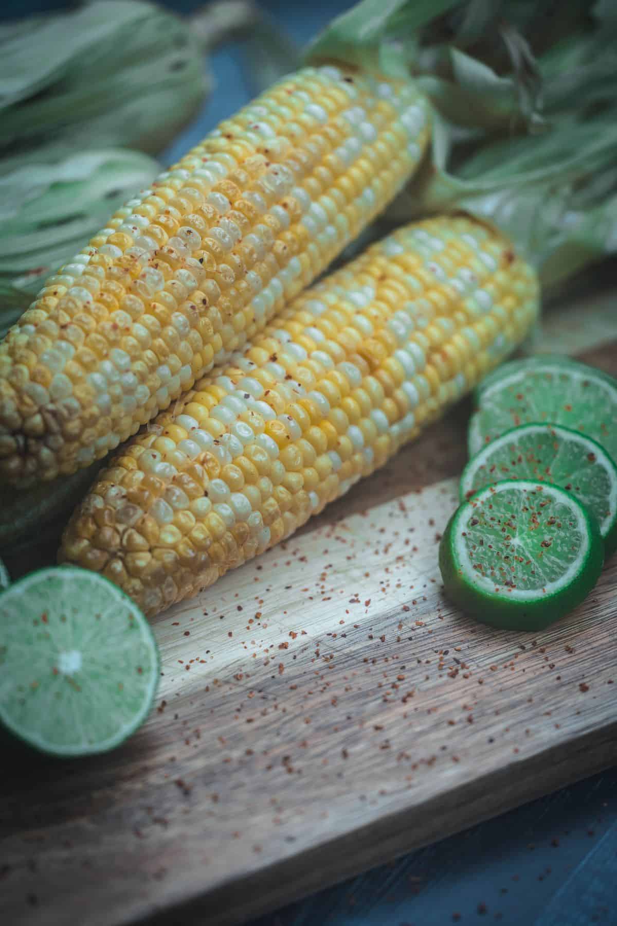 Vertical photo of smoked corn on the cob with chili lime butter, fresh lime slices, and red pepper flakes on a wooden cutting board.