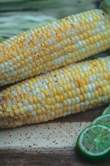 Smoked corn on the cob brushed with chili lime butter, served with fresh lime slices and red pepper flakes on a wooden board.
