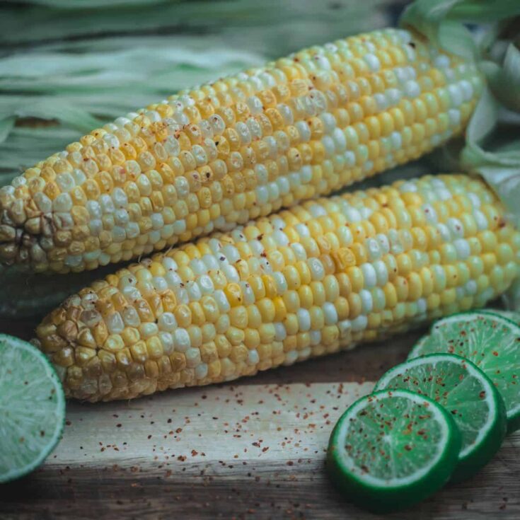 Smoked corn on the cob brushed with chili lime butter, served with fresh lime slices and red pepper flakes on a wooden board.