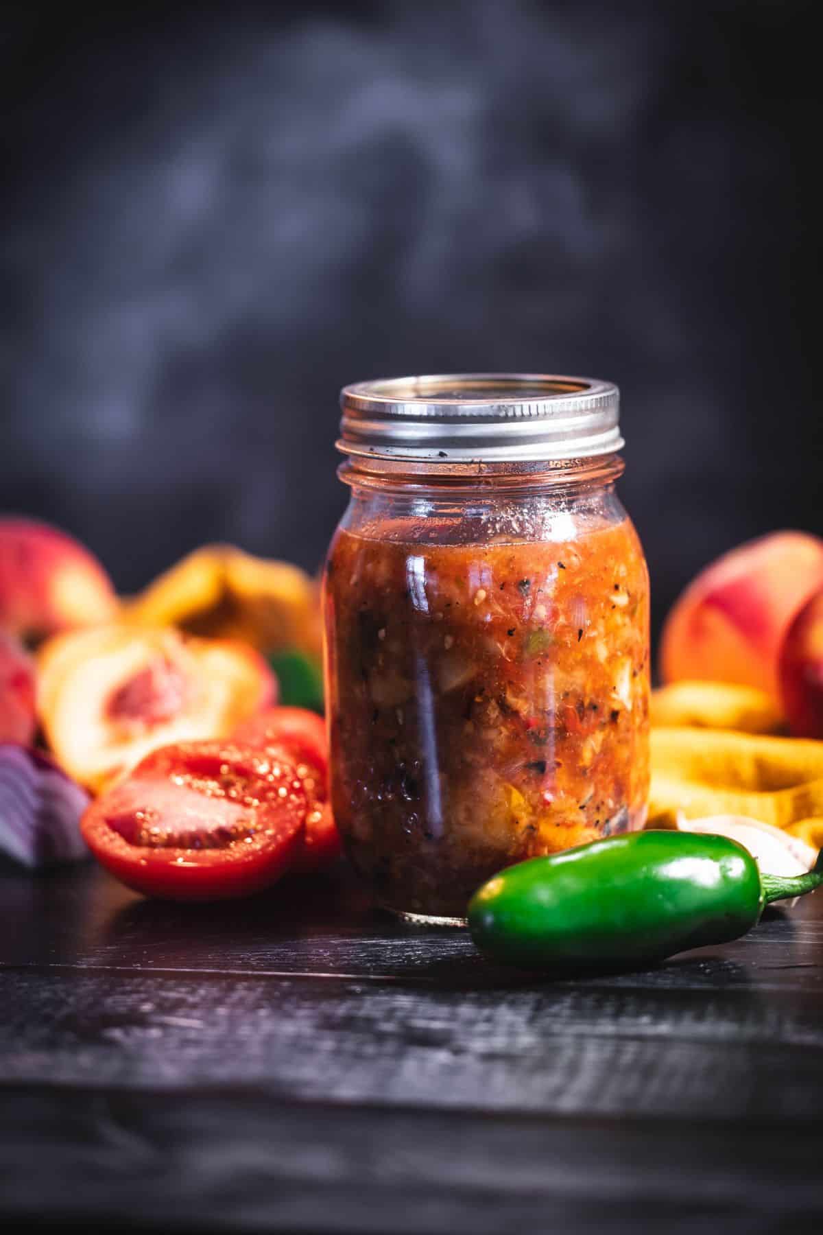 Jar of roasted peach salsa with fresh peaches, tomato, red onion, garlic, and jalapeño on a dark wooden surface.
