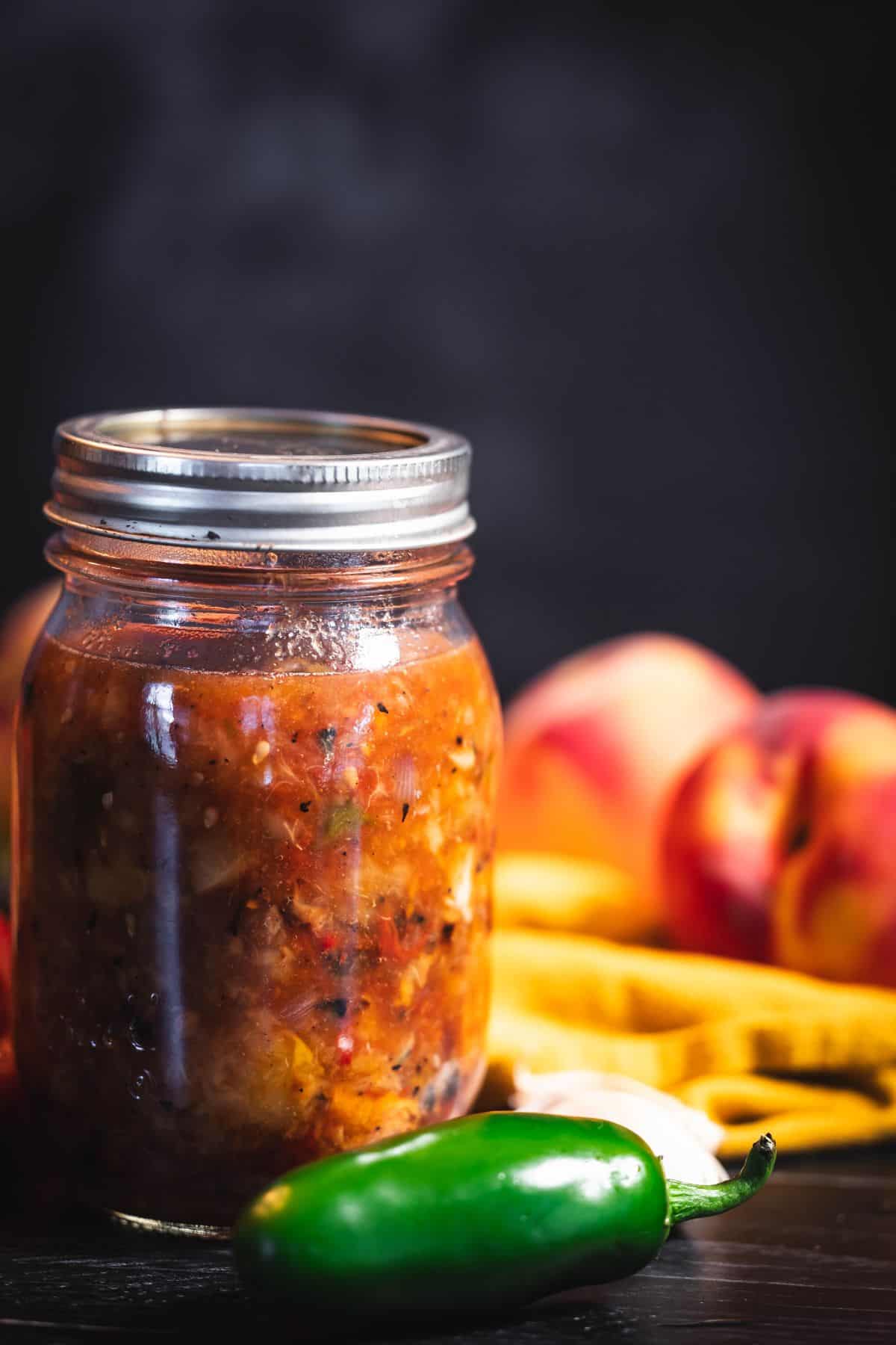 Jar of roasted peach salsa with a jalapeño, garlic, and fresh peaches on a dark surface.