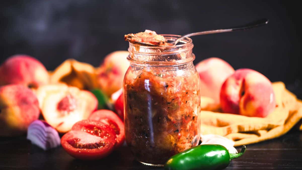 Spoonful of chunky roasted peach salsa over an open jar, surrounded by fresh peaches, tomatoes, red onion, garlic, and jalapeño.