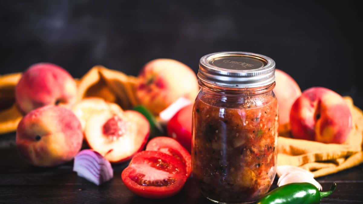 Jar of roasted peach salsa with fresh peaches, tomatoes, red onion, garlic, and jalapeño on a dark surface.